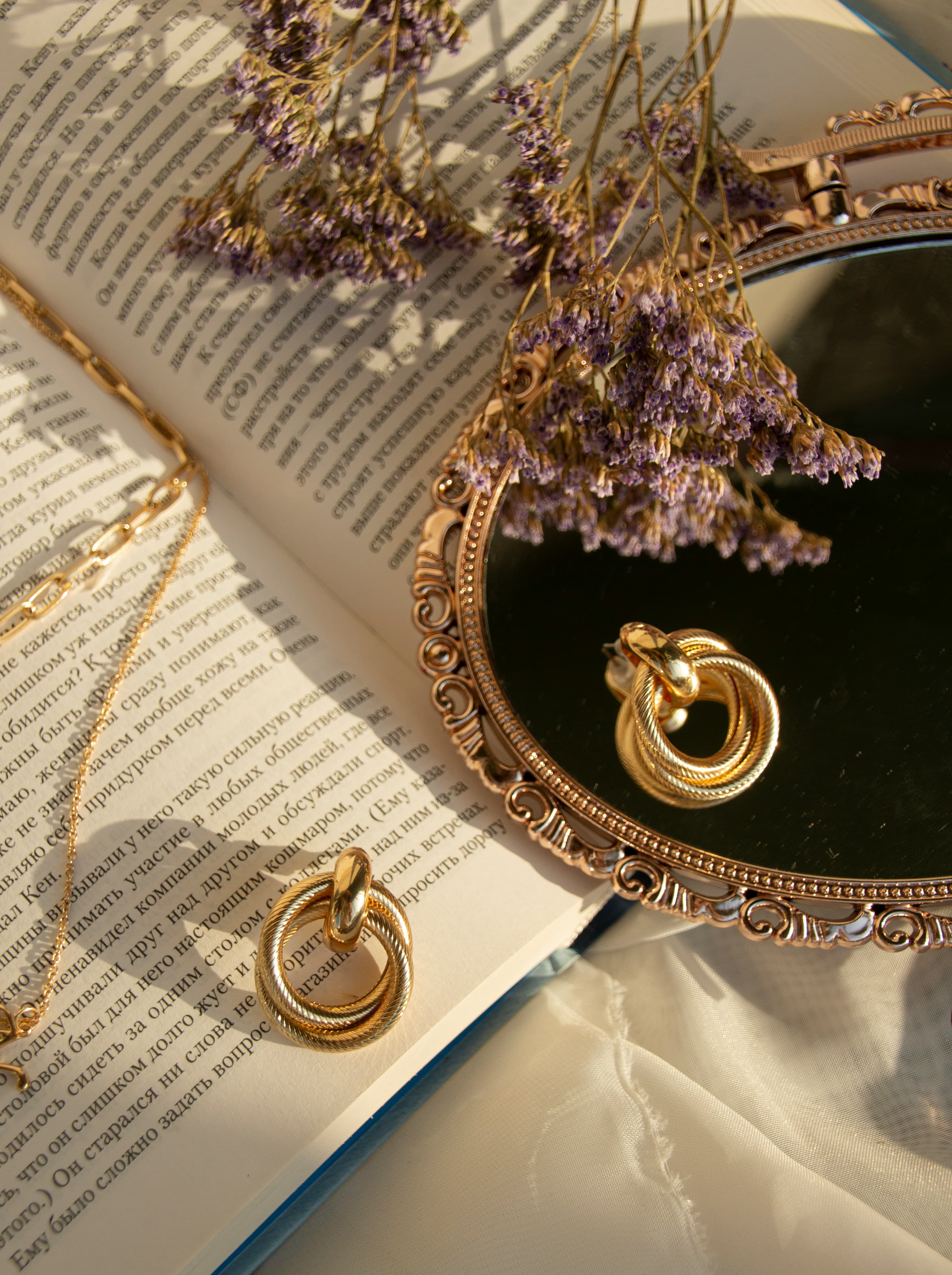 Gold hoop earrings and chain necklace resting on an open book beside an ornate mirror, with dried lavender